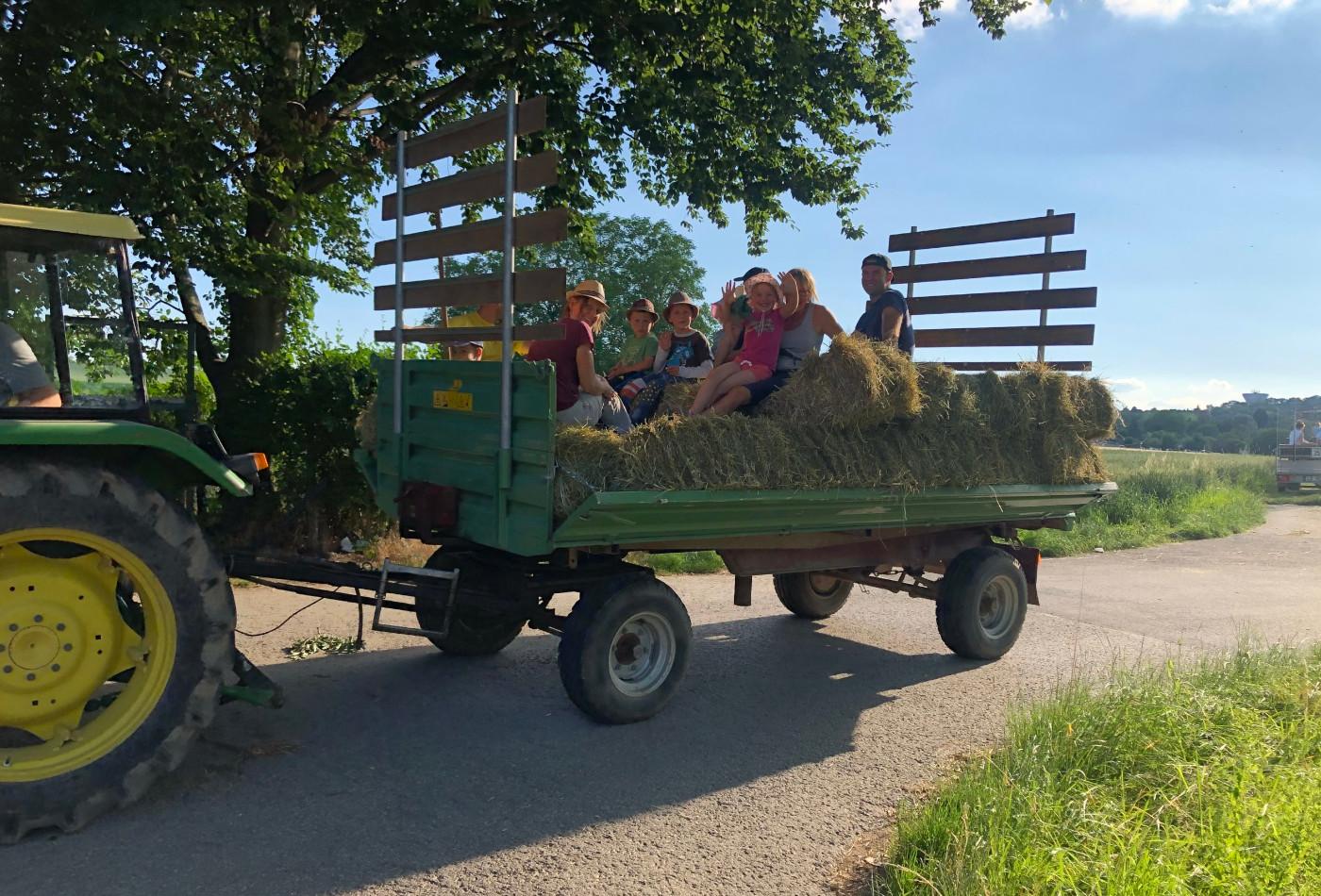 Kinder sitzen winkend auf einem mit Heu beladenen Traktor-Anhänger