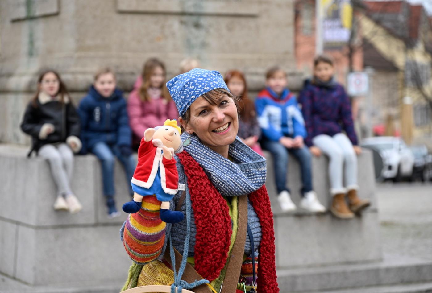 Stadtführerin im Kostüm mit Handpuppe, im Hintergrund sitzen mehrere Kinder auf dem Obelisken am Kaffeeberg in Ludwigsburg