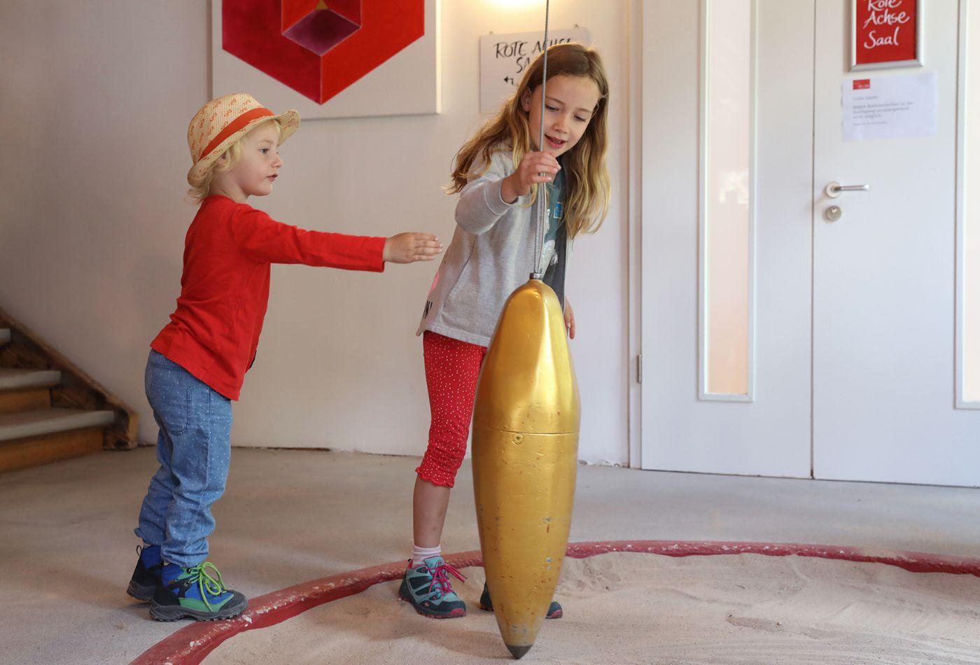 Ein Mädchen und ein Junge bewegen einen am Band hängenden Kegel, dessen Spitze im Sand Linien zieht.