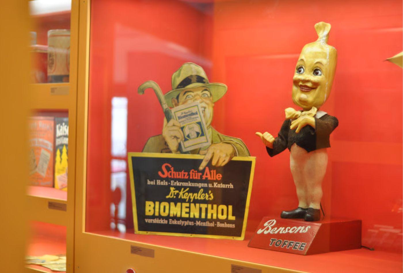 Neben einem alte Bonbon-Reklameschild steht eine Figur, deren Kopf ein Bonbon ist