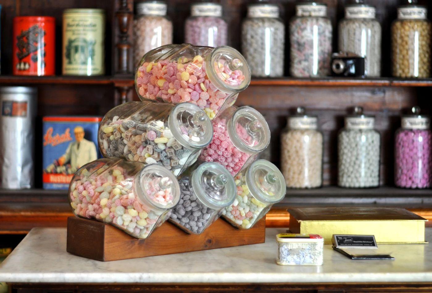 Pyramidenförmig gestapelte Bonbongläser mit bunten Bonbons stehen auf einem Tresen vor einem Regal, in dem weitere Bonbongläser stehen.