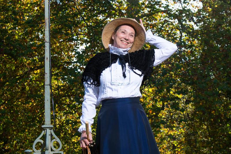 Eine Frau in langem, blauen Rock, weißer Bluse und einem schwarzen Schal um den Schultern hält ihren Stohhut fest und lacht.