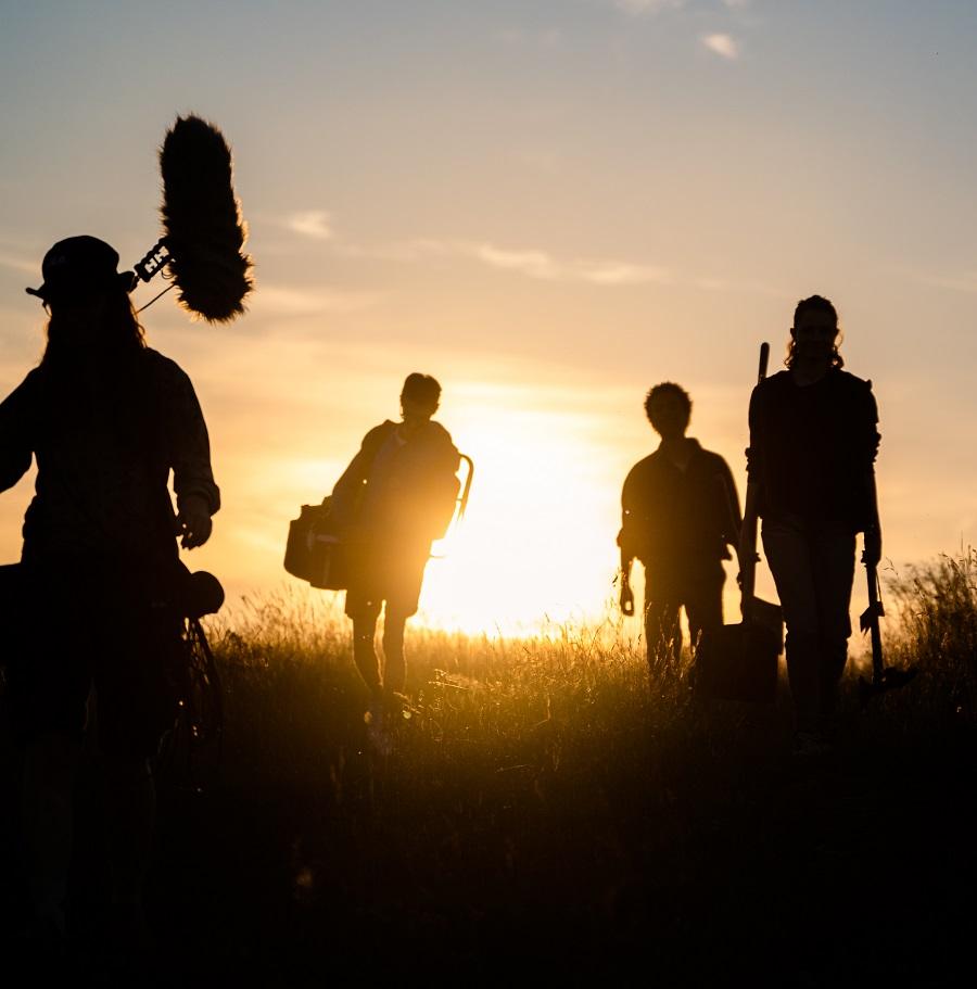 Es ist ein Filmteam zu sehen, welches bepackt mit ihrem Equipment auf einer Wiese in den Sonnenuntergang läuft.