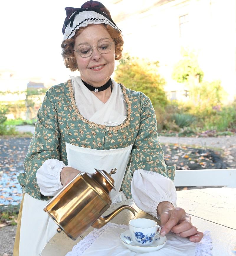 Eine historisch gekleidete Frau gießt aus einer goldenen Kanne ein Getränk in eine kleine Porzellantasse