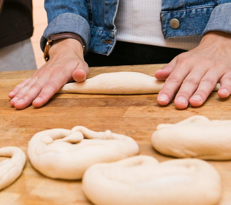 Man sieht die Hände einer Frau, die Brezelteig ausrollt und ein paar fertige Brezel-Rohlinge auf einem Tisch