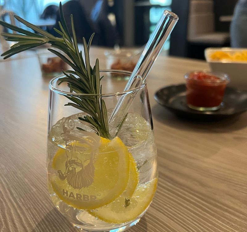 Das Foto zeigt einen Longdrink-Glas mit einem eisgekühlten Getränk mit Zitronenscheiben, einem gläsernen Trinkhalm und einem kleinen Rosmarinzweig