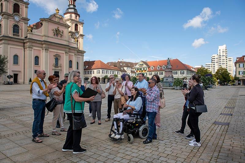Eine Gruppe von Menschen steht vor der Stadtkirche auf dem Marktplatz und hört gespannt einer Gästeführerin zu.