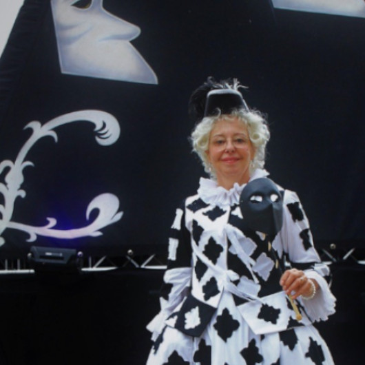 Harlekina blickt mit hellen Locken und Brille in die Kamera. Sie trägt ein schwarz-weiß gemustertes Kleid und einen schwarzen Hut mit Feder.