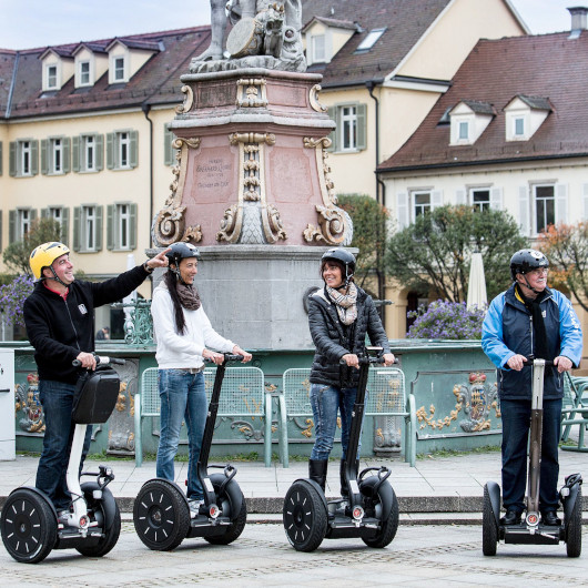 Vier Menschen stehen auf Segways, im Hintergrund sieht man den Brunnen auf dem Marktplatz.