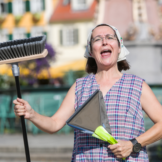 Eine Frau mit Besen und Kehrschaufel in einer Kittelschürze und Tuch auf dem Kopf schaut lustig in die Kamera.