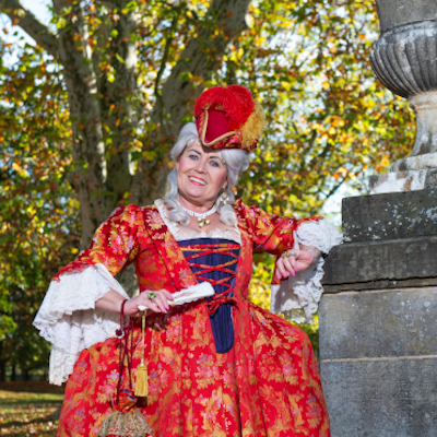 Reichsgräfin Wilhelmine von Grävenitz steht in einem imposanten, roten Kleid vor einer Statue. Sie blickt freudig in die Kamera.