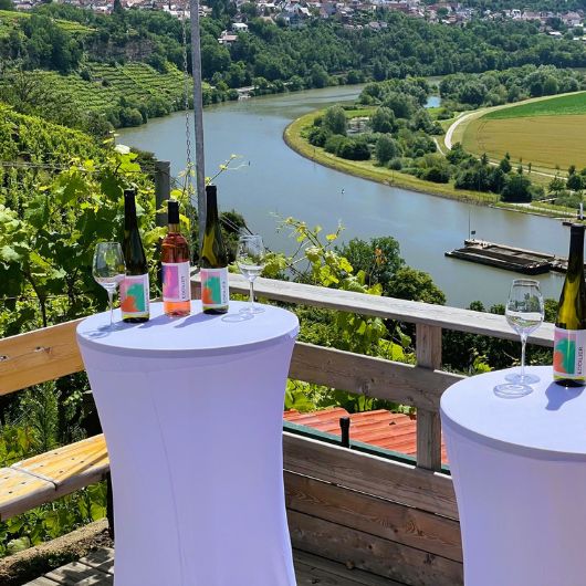 Zwei Stehtische mit weißen Hussen, auf denen verschiedene Weine stehen. Im Hintergrund Weinberge und der Neckar