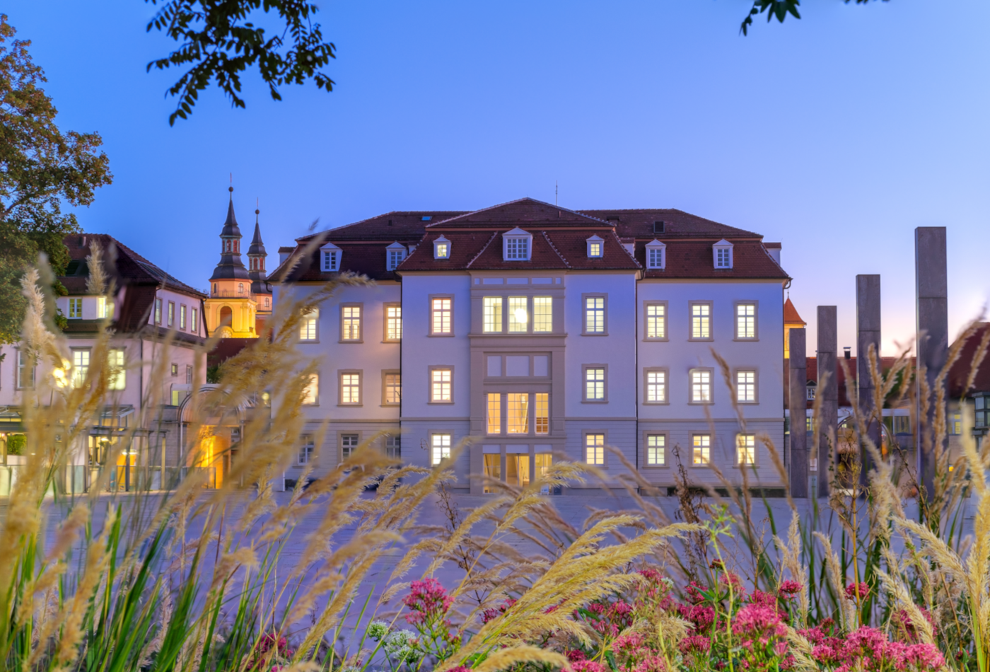 Das Rathaus bei Dämmerung, im Vordergrund Blumen und Gräser, im Hintergrund das Rathaus mit leuchtenden Fenstern.