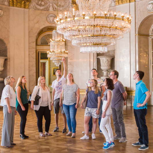 Zu sehen ist eine Gruppe, die eine Führung im Residenzschloss besucht - sie bestaunen den über ihnen hängenden Kronleuchter.