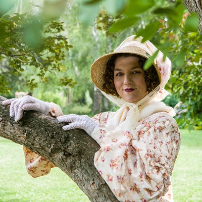 Eine Frau mit Hut, Spitzenhandschuhen und Blumenkleid lehnt an einem Baum auf einer Wiese.