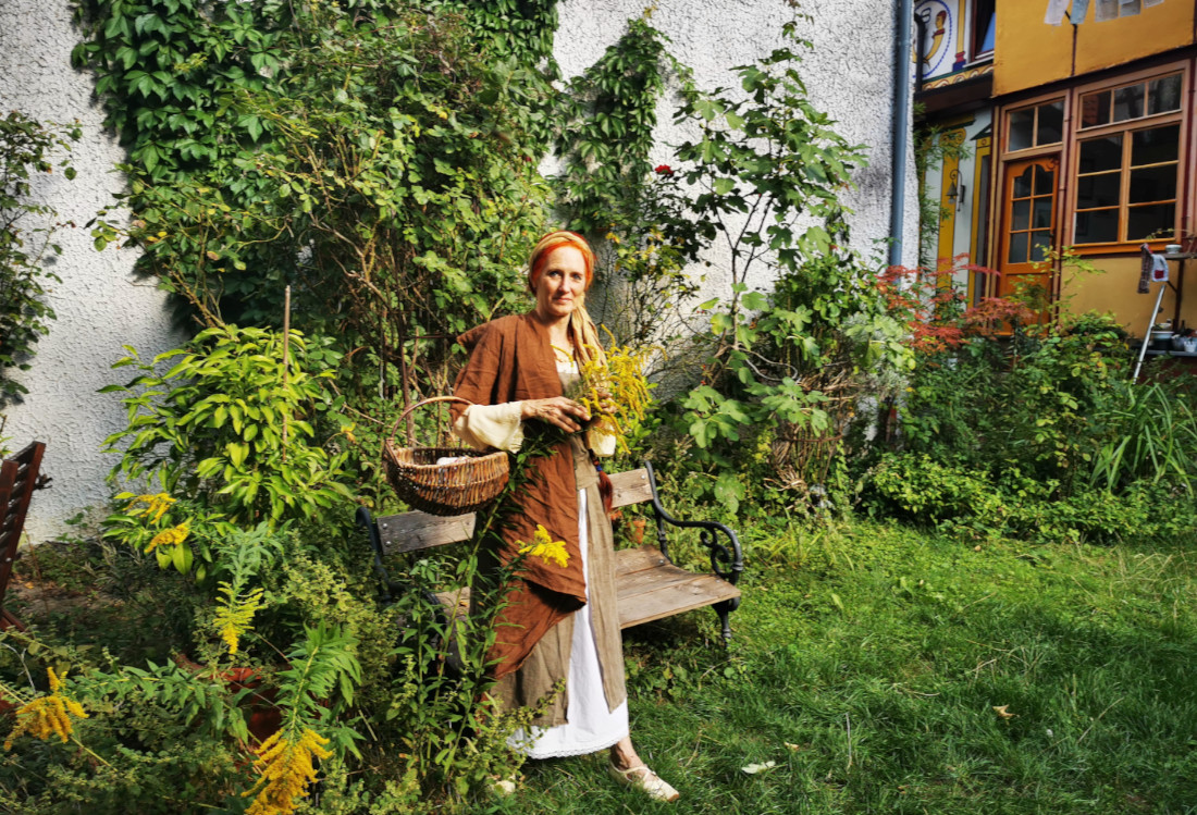 Agatha von Schönhausen Eine kostümierte Frau steht auf einer Wiese, trägt einen Korb und hält Kräuter in der Hand.
