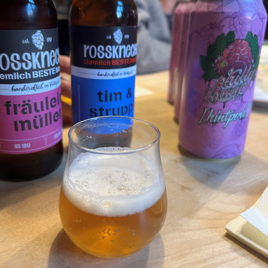 Ein gefülltes Bierglas steht auf einem Tisch, dahinter zwei Bierflaschen und Dosen