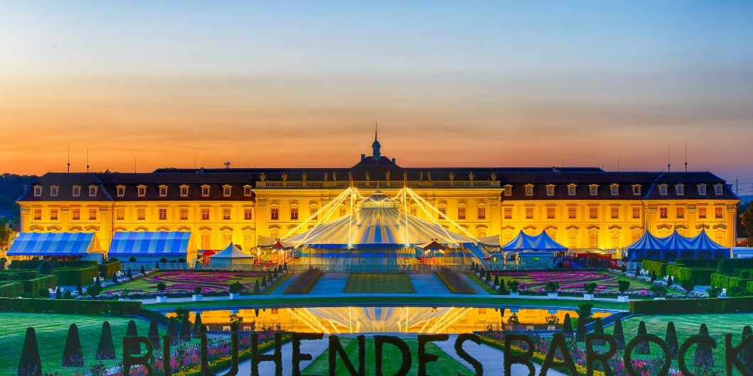 Blick auf das beleuchtete Zirkuszelt und die ebenfalls beleuchtete Fassade des Residenzschlosses Ludwigsburg bei Sonnenuntergang.