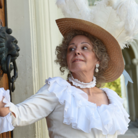Eine Frau mit weißem Gewand und Hut klopft an einer Tür.
