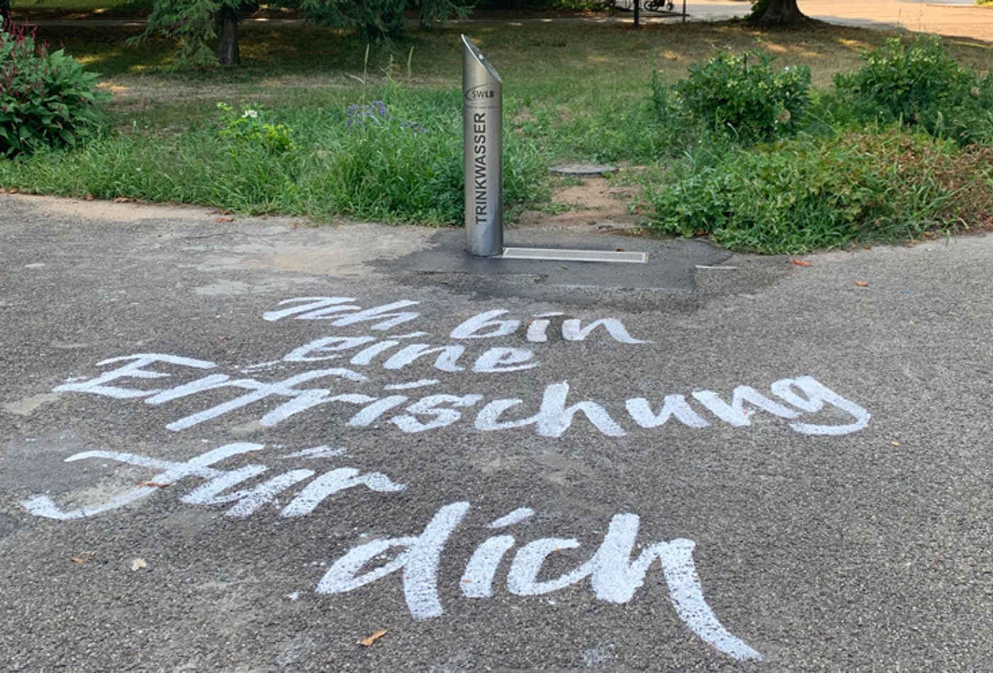 Blick auf einen Trinkwasserbrunnen. Auf dem Asphalt vor dem Brunnen steht in weißer Schrift: "Ich bin eine Erfrischung für dich"