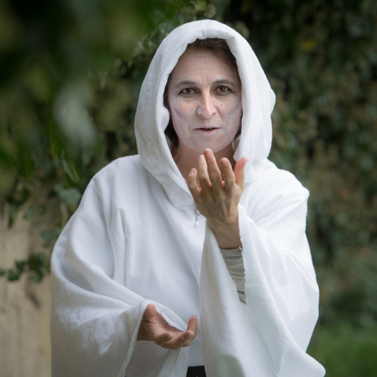 Eine blass geschminkte Frau lockt mit einer Handbewegung zu sich. Sie trägt ein weißes Gewand mit Kapuze.