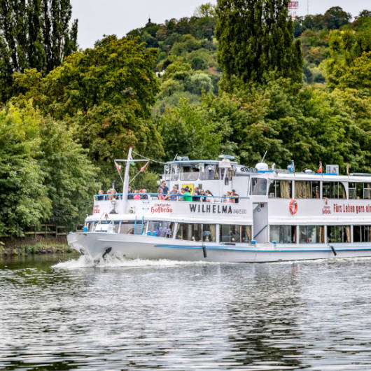 Weißes, zweistöckiges Ausflugsschiff auf einem Fluss im Grünen.