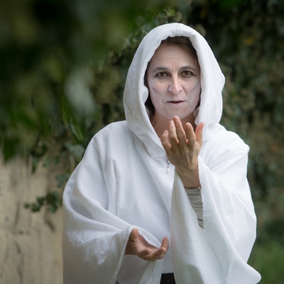 Eine blassgeschminkte Frau lockt einen mit einer Handbewegung zu sich. Sie trägt ein weißes Gewand mit Kapuze.