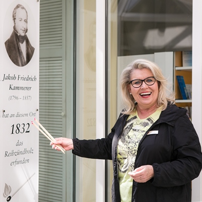 Eine Frau mit einer Brille und Streichhölzern steht vor einem Bild von Jakob Friedrich Kammerer