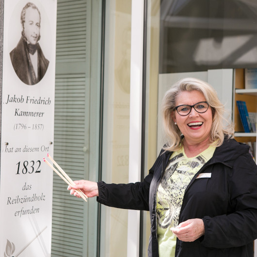 Eine Frau mit einer Brille und Streichhölzern in der Hand steht vor einem Bild von Jakob Friedrich Kammerer.