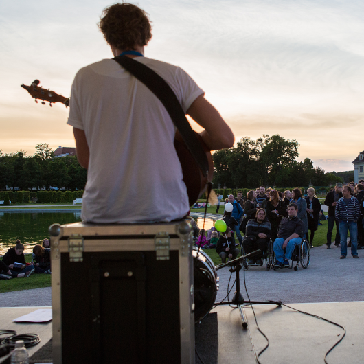 Zu sehen ist ein Musiker mit Gitarre von hinten, welcher einem kleinen Publikum vorspielt.