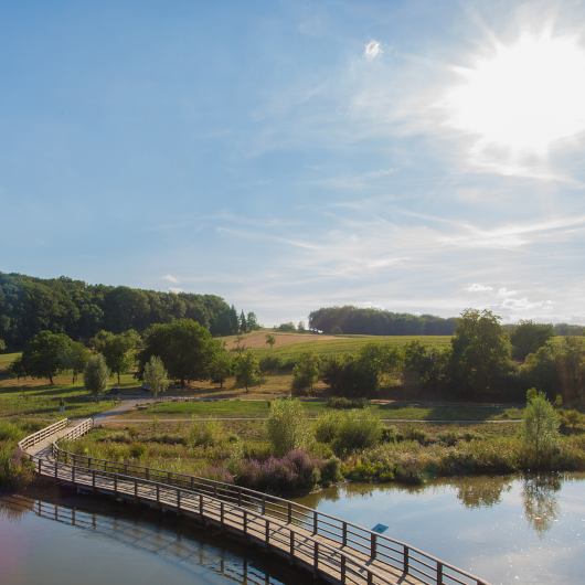 Zu sehen ist ein Fluss mit einer Brücke, im Hintergrund sind Wiesen und Wälder erkennbar.