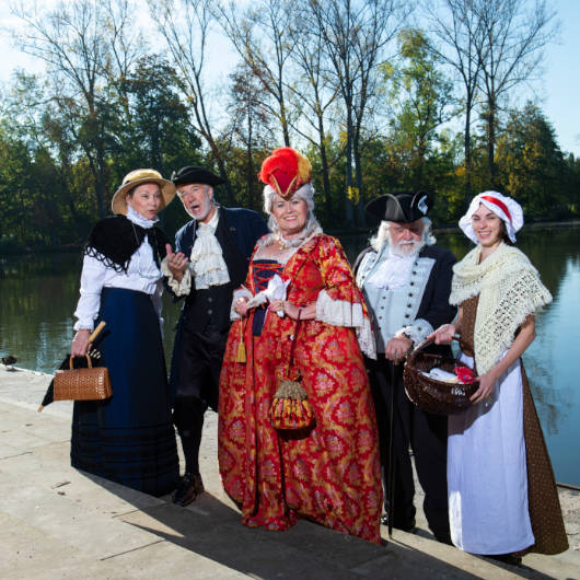 Fünf in historische Gewänder gekleidete Personen stehen vor einem See.