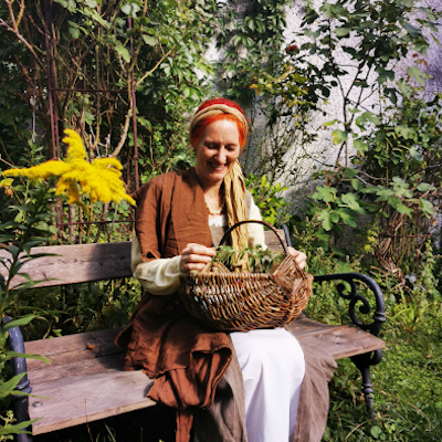 Die Kräuterhexe Agathe sitzt auf einer Holzbank, sie hat einen geflochtenen Korb auf dem Schoß und schaut auf die Pflanzen  in ihrer Hand.