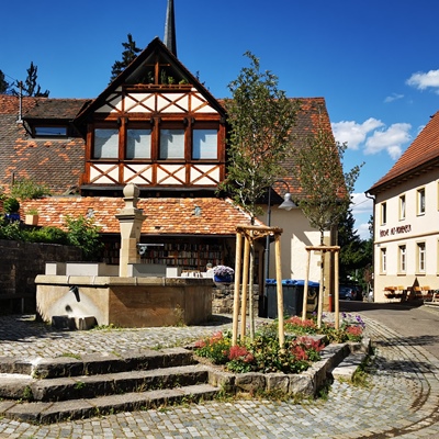 Blick auf den Stadtteil Alt-Hoheneck. Im Vordergrund ein von Bäumen umrundeter Brunnen, im Hintergrund ein Fachwerkhaus.