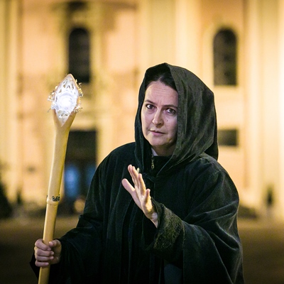 Eine Frau, blass geschminkt, in einem schwarzen Gewand mit Kapuze und einem leuchtenden Stab blickt in die Kamera
