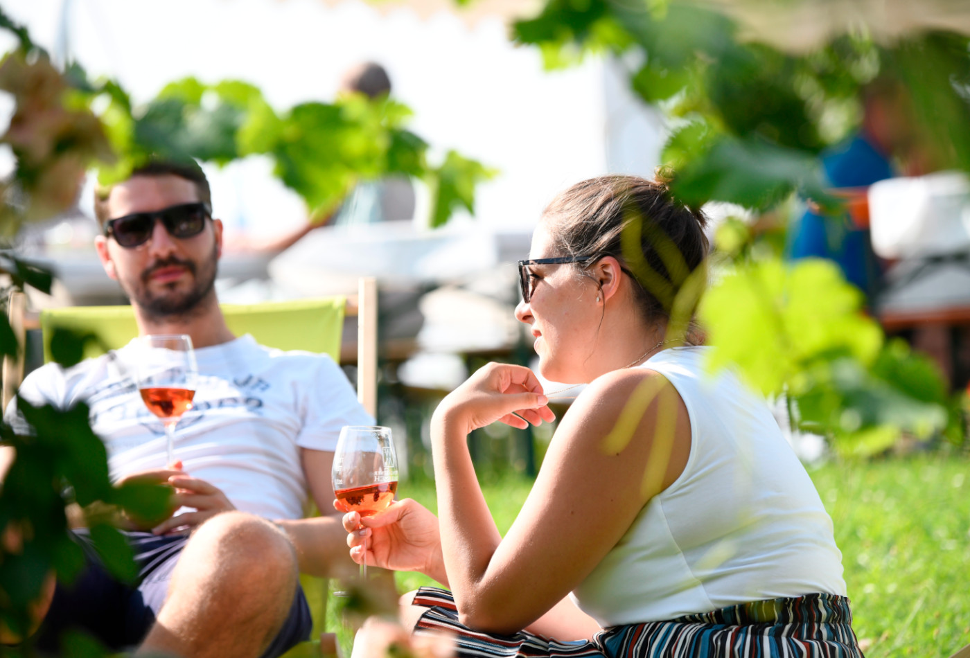 Zwei Personen sitzen in Liegenstühlen mit Weingläsern in der Hand in einer grünen Umgebung.