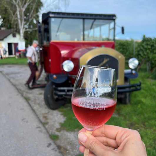 Man sieht eine Hand, die ein Glas Rotwein hält, im Hintergrund ein roter Oldie-Bus und Weinberge