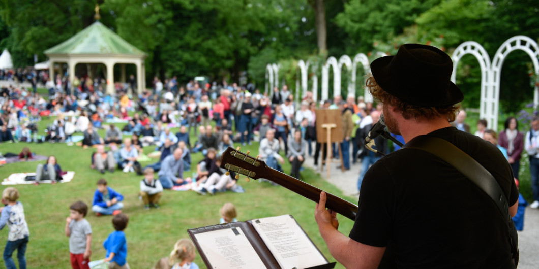 Im Vordergrund sieht man einen Gitarrenspieler von hinten, im Hintergrund eine Wiese, auf der viele Menschen sitzen und zuhören.