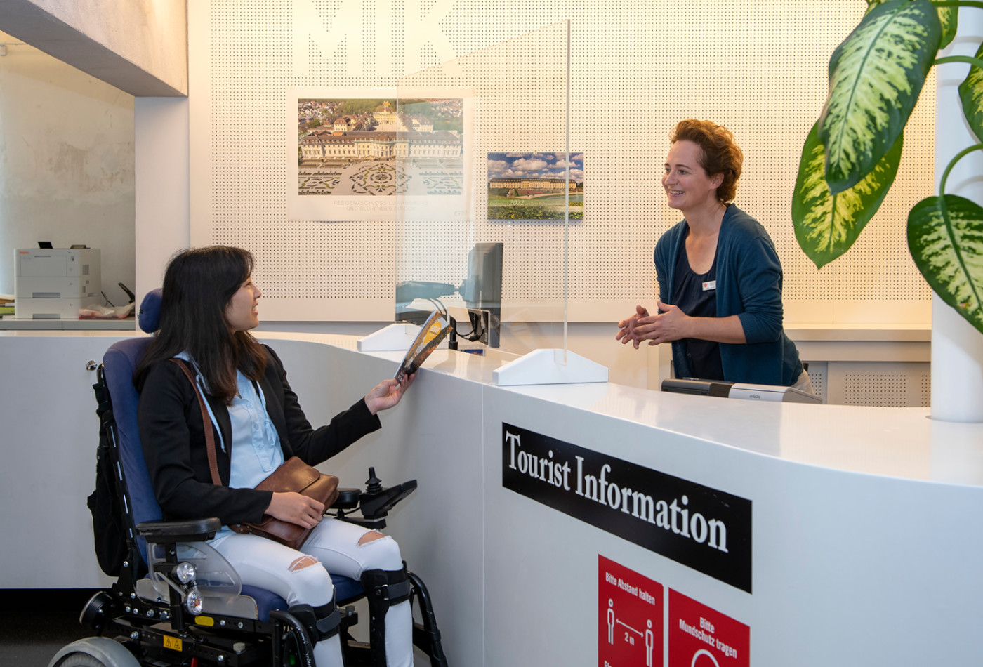 Frau im Rollstuhl lässt sich von einer Mitarbeiterin in der Tourist Info beraten.
