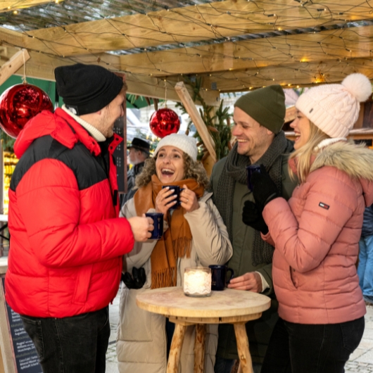 Zu sehen sind vier Personen, die an eine Tisch zusammenstehen und Glühwein trinken