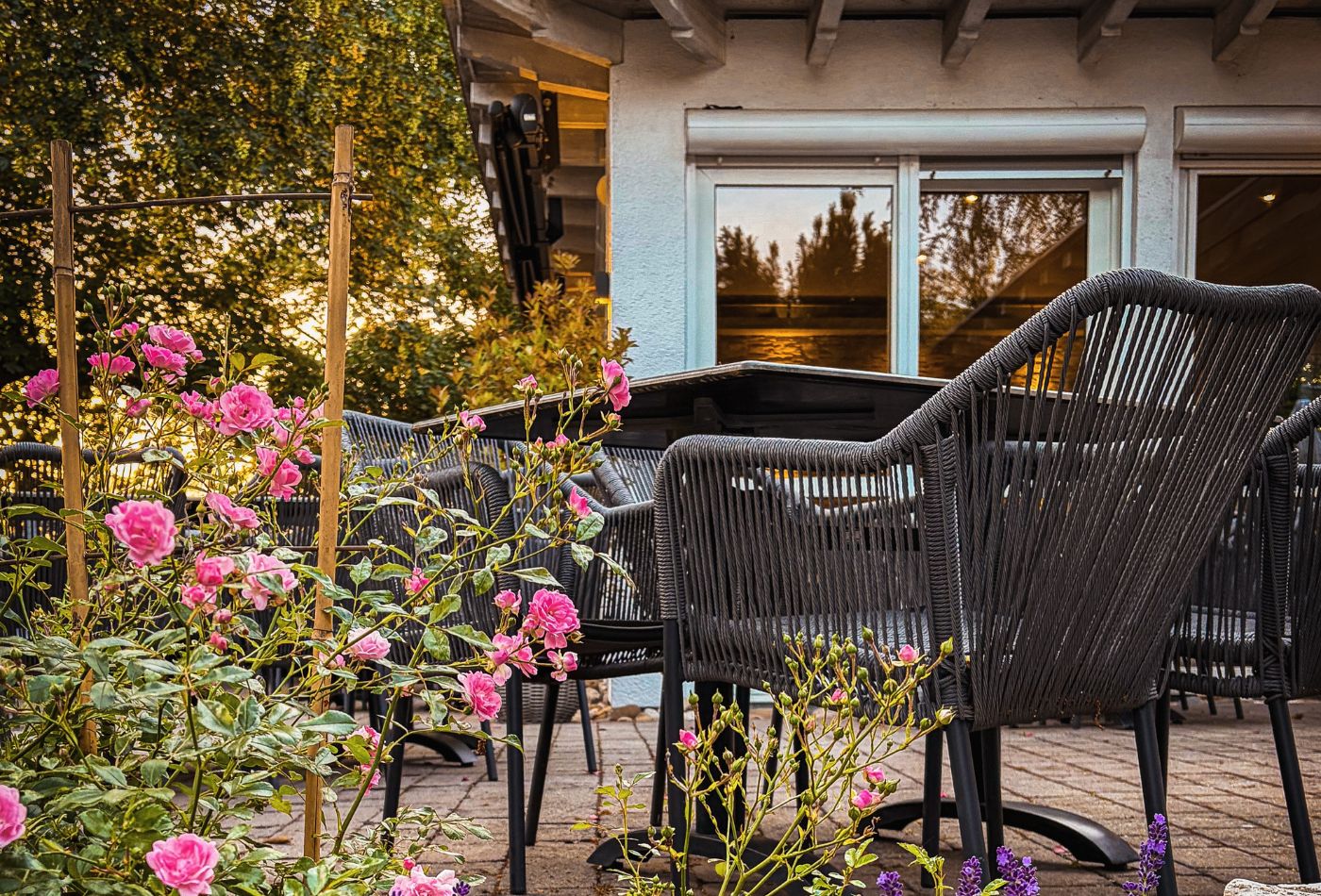 Blick auf eine Terrasse, im Vordergrund blühen rosafarbene Rosen, im Hintergrund sieht man ein Haus
