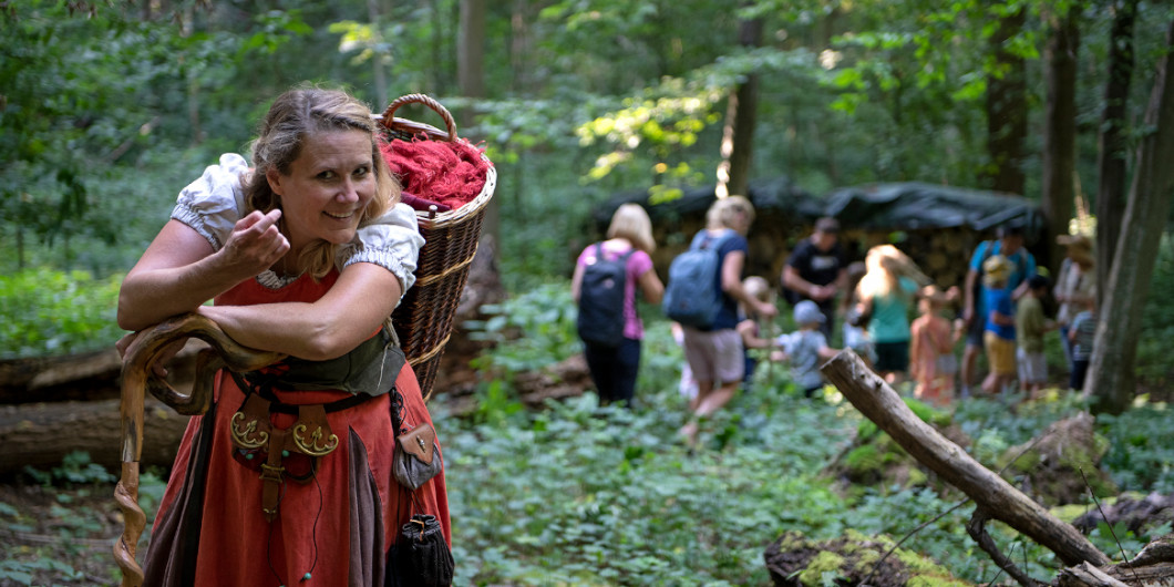Eine Gästeführerin in alterümlichem Gewand ist mit einer Gruppe Kindern in der Natur unterwegs.