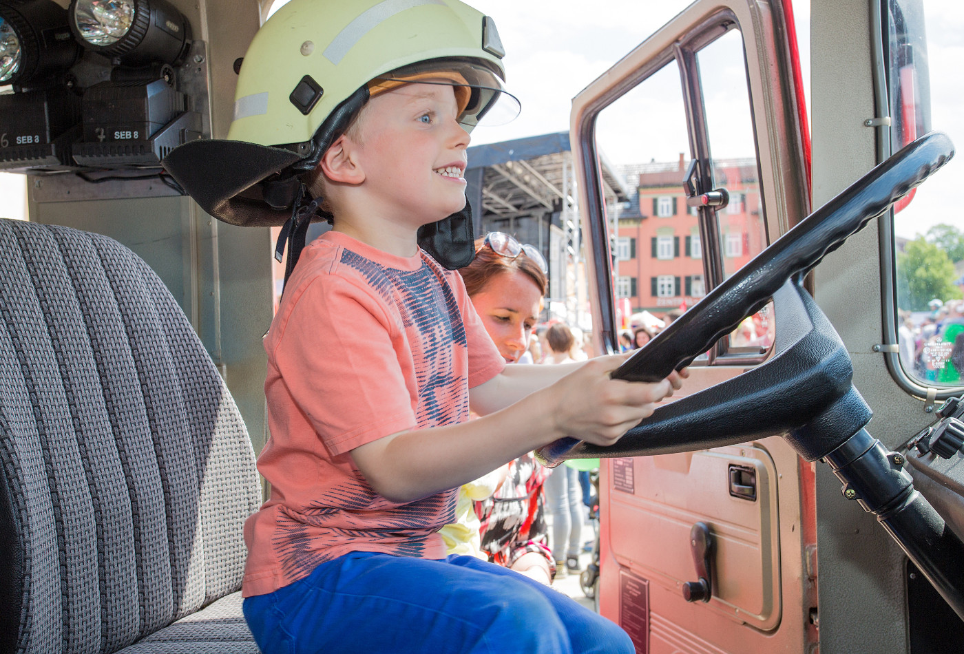 Ein Kind sitzt grinsend am Steuer eines Feuerwehrautos und hat einen Feuerwehrhelm auf