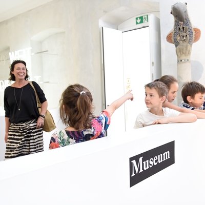Vier Kinder sind mit einer erwachsenen Person im Museum und schauen sich Exponate an.