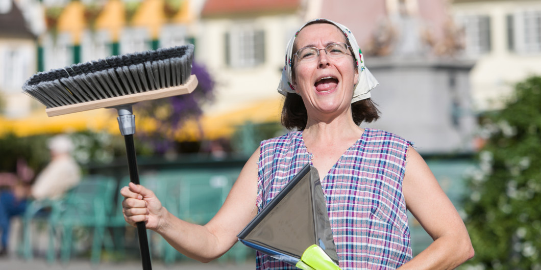Eine Frau mit Besen und Kehrschaufel in einer Kittelschürze und Tuch auf dem Kopf schaut lustig in die Kamera.