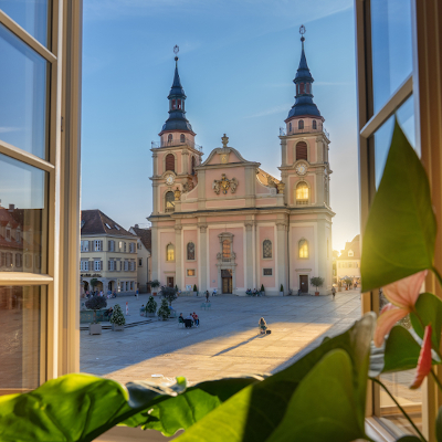 Die evangelische Stadtkirche, buntes Treiben auf dem Marktplatz, Ansicht durch ein Fenster mit Pflanzen, ein Sonnenstrahl fällt ins Bild