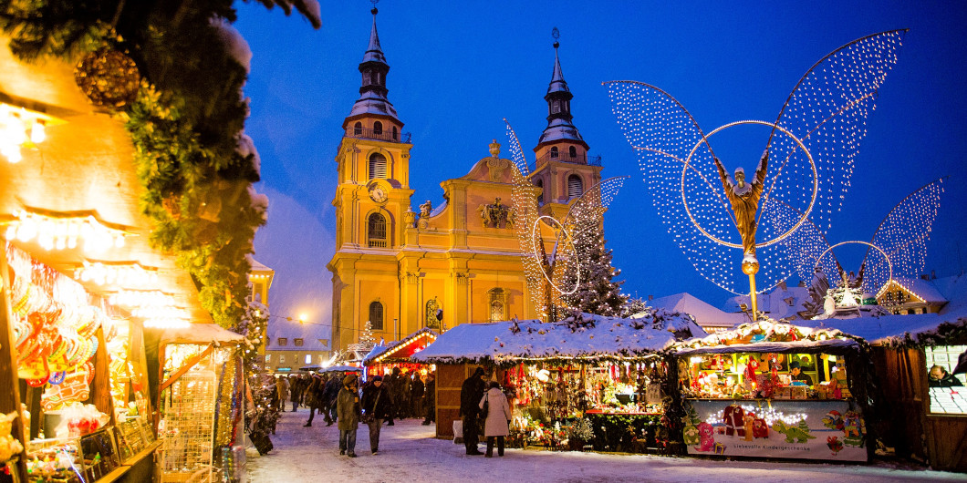 Blick auf den Weihnachtsmarkt Ludwigsburg zur blauen Stunde. Man sieht schneebedeckte Stände, die leuchtenden Engelsfiguren und die beleuchtete evangelische Kirche