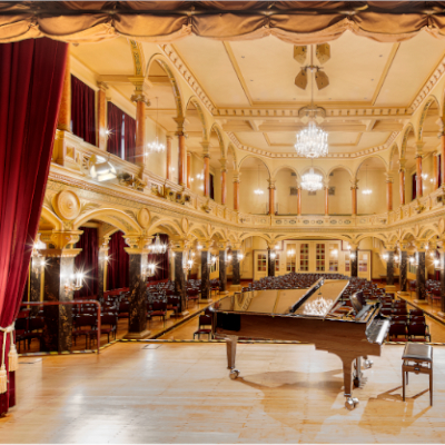 Innenansicht der Musikhalle. Man blickt von der Bühne aus, auf der ein Klavier steht in den Publikumssaal, der leer ist.