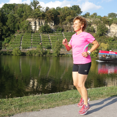 Eine Frau in Sportkleidung joggt am Neckarufer entlang, im Hintergrund sieht man Weinberge und ein Boot