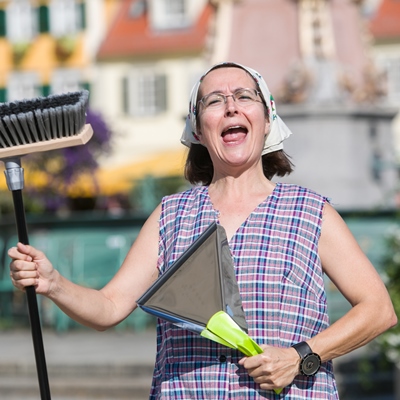 Eine Frau mit Besen und Kehrschaufel in einer Kittelschürze und Tuch auf dem Kopf schaut lustig in die Kamera